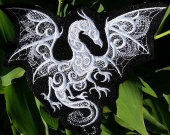 Puede incluir: Parche de tela negro con un diseño de dragón bordado en blanco. El dragón está enroscado con las alas extendidas.
