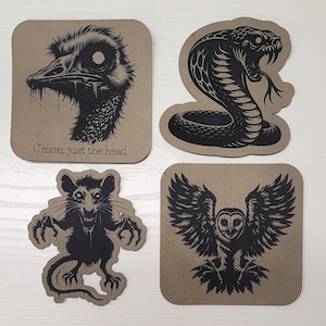 May include: Set of four coasters featuring black and white illustrations of an ostrich head, a cobra, a rat, and an owl. The coasters are made of brown leather-like material and have a textured surface. The ostrich coaster has the text "C'mon, just the head." printed below the illustration.