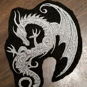 May include: A black and white embroidered patch featuring a dragon with its wings spread out. The dragon is outlined in white thread and the background is black.