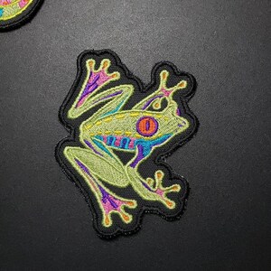 May include: Embroidered patch featuring a stylized tree frog. The frog has a light green body with blue, purple, and orange accents. The patch is outlined in black.
