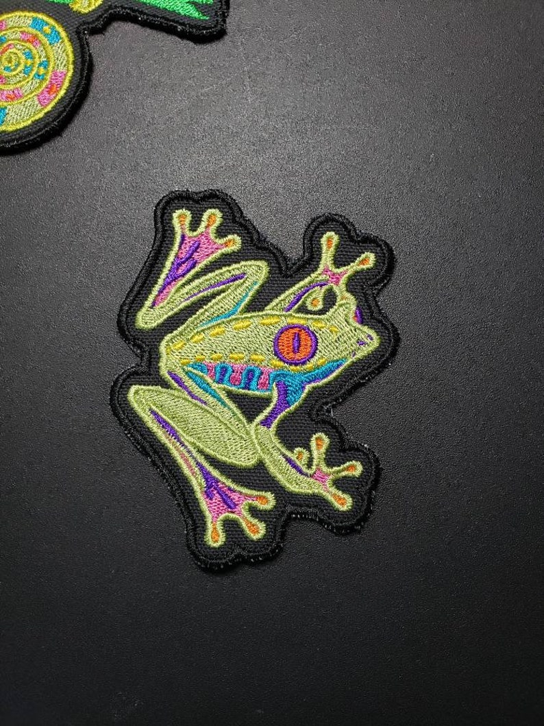 May include: Embroidered patch featuring a stylized tree frog. The frog has a light green body with blue, purple, and orange accents. The patch is outlined in black.