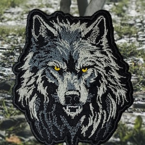May include: Embroidered patch of a wolf's face with a black background and white fur. The wolf has yellow eyes and a fierce expression.