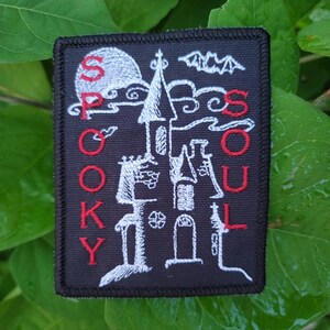 May include: A black embroidered patch with a white silhouette of a spooky house and a bat. The words "Spooky Soul" are embroidered in red on the left and right sides of the patch.