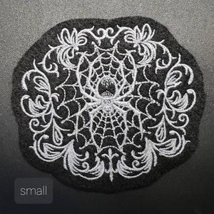May include: A black embroidered patch with a white spider web and spider in the center. The spider web is surrounded by a white floral design.