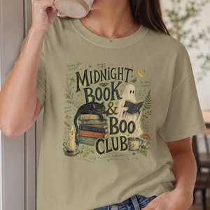 Könnte beinhalten: Ein beiges Kurzarm-T-Shirt mit einem Grafikdruck und dem Text "THE MIDNIGHT BOOK & BOO CLUB". Das Design zeigt einen Bücherstapel, eine schwarze Katze, ein lesendes Gespenst, eine brennende Kerze, eine Tasse und botanische Elemente in Grün und Braun.