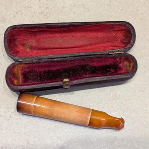 Könnte beinhalten: Eine Vintage-Tabakpfeife mit einem hellbraunen Holzkörper und einem dunkelbernsteinfarbenen Mundstück liegt neben einem offenen, dunkelvioletten Etui mit rotem Samtfutter. Das Etui ist rechteckig mit einem goldenen Verschluss.