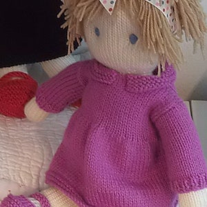 May include: A knitted doll with blonde yarn hair wearing a pink dress and a headband with a polka dot bow. The doll is sitting on a white surface.