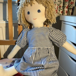 May include: A handmade rag doll with blonde yarn hair, blue eyes, and a white dress with blue stripes. The doll is sitting on a chair with a floral pattern.