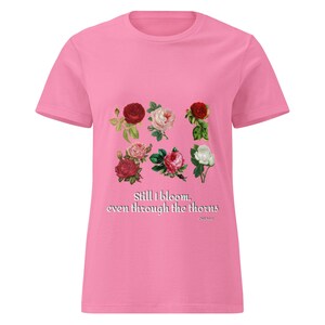 Könnte beinhalten: Rosafarbenes T-Shirt mit einem Blumendesign aus neun Rosen in Rot-, Rosa- und Weißtönen. Der Text "Still I bloom, even through the thorns" ist darunter gedruckt. Ein lässiges Oberteil mit kurzen Ärmeln.