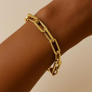 May include: A gold-colored chain link bracelet. The bracelet is made of rectangular links and is worn on a wrist. The links are connected to each other to form a continuous chain. The bracelet is a bright, shiny gold color.