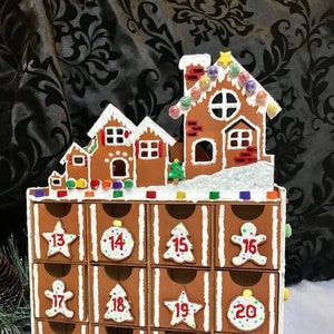 May include: A gingerbread house shaped advent calendar with 24 drawers. Each drawer has a number from 13 to 24 and a festive design, including a star, a tree, a gingerbread man, and an ornament.