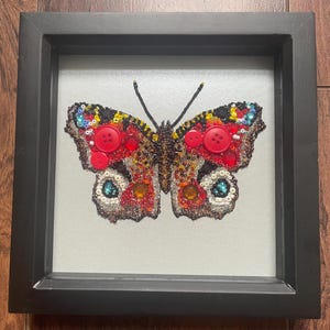 Op de afbeelding: Een ingelijst vlinderkunstwerk. De vlinder is gemaakt met kralen en knopen in rood, bruin, geel en blauw. Het kunstwerk is ingekaderd in een zwart frame, wat een contrast creëert. De vleugels van de vlinder zijn gedetailleerd.