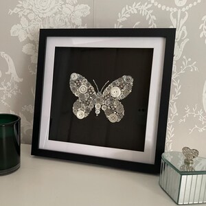 Könnte beinhalten: Gerahmtes Kunstwerk mit einem Schmetterling aus grauen und weißen Knöpfen auf schwarzem Hintergrund. Das Kunstwerk befindet sich in einem schwarzen Rahmen, ausgestellt auf einer weißen Oberfläche, mit einem grünen Glas-Kerzenhalter und einer verspiegelten Schmuckschatulle.