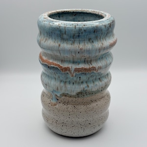 May include: A handmade ceramic vase with a unique, textured design. The vase features a light blue and brown glaze over a speckled, off-white base. The vase has a cylindrical shape with a wavy, ribbed texture.