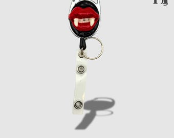 Pode incluir: Um porta-crachá retrátil com um design de boca de vampiro. O porta-crachá apresenta uma boca vermelha com presas brancas, um fundo preto e um clipe prateado. Uma alça branca com dois encaixes e um anel de metal estão presos. O texto "SLICE OF HEAVEN" está na parte inferior.