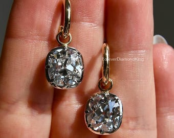 May include: A pair of gold hoop earrings featuring square, clear gemstones. The earrings are held in a hand, highlighting the sparkling stones and gold setting. A classic jewellery design.