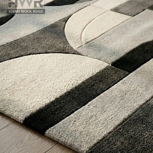 Pode incluir: Close-up de um tapete de lã tufado à mão com um design geométrico. O tapete apresenta um padrão de linhas curvas e retas em tons de cinza, preto e branco sujo. O texto "HWR HAND WOOL RUGS" está no canto superior esquerdo.