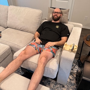 May include: A person wearing a black t-shirt and floral print shorts is seated on a light-colored sectional sofa. The sofa has a built-in side table with a wooden phone holder. A footrest is extended.