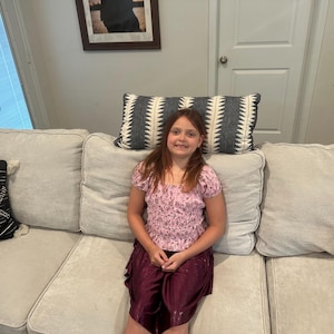 May include: A young person wearing a pink floral top and burgundy skirt sits on a light gray sofa. Decorative pillows with geometric patterns in black and white are visible. An orange pillow is on the right side of the sofa.