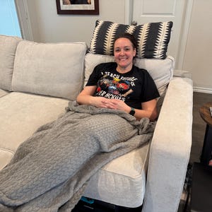 May include: A woman is seated on a light gray sofa, covered with a gray blanket. She wears a black t-shirt with the text "I TEACH FUTURE SUPER HEROES". Decorative pillows with black and white patterns are behind her.
