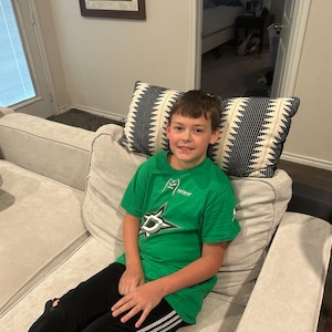 May include: A person wearing a green t-shirt with a Dallas Stars logo, black pants with white stripes, sitting on a light gray couch. Two decorative pillows with black and white patterns are behind the person.