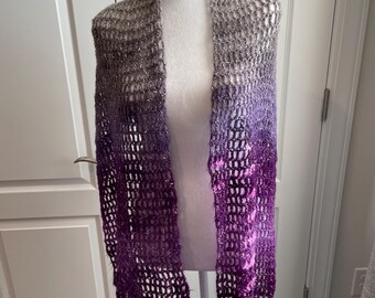 May include: A handmade, open-weave shawl in a gradient of colors, from silver to purple. The shawl is draped over a mannequin, showcasing its texture and design. The open weave creates a delicate, airy look.
