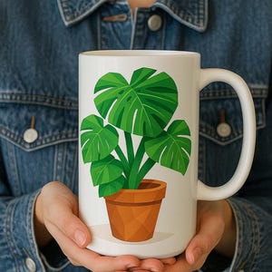 May include: White ceramic mug featuring a green illustration of a potted Monstera plant. The plant has large, split leaves and is in a brown pot. The mug has a curved handle.