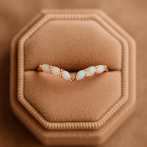 May include: A gold ring with a V-shaped design, set with oval opals. The ring is presented in a brown velvet jewellery box. The opals are milky white with a subtle iridescent sheen.