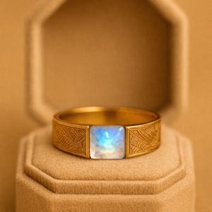 May include: A gold ring with a rectangular, iridescent gemstone center. The band features an etched design. The ring is displayed on a beige, octagonal jewelry box, set against a warm, neutral background. A piece of jewelry.