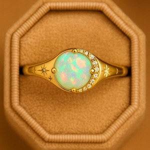 May include: A gold ring with a large, round, iridescent opal center stone. The ring features a halo of small, clear gemstones and starburst and line designs on the band. The ring is displayed in a brown ring box.