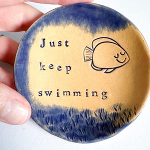 Puede incluir: Un plato redondo de cerámica con un centro beige y un borde azul. El plato presenta las palabras "Just keep swimming" y una ilustración de un pez sonriente. El borde inferior tiene un diseño de olas azules.