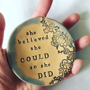 Può includere: Un sottobicchiere rotondo in ceramica con sfondo marrone chiaro e bordo blu. Il sottobicchiere presenta un motivo floreale e il testo "she believed she could so she did" in nero.