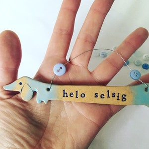 Op de afbeelding: Een keramisch teckelbeeldje met een blauwe en gele glazuur. Het beeldje houdt een bord vast met de tekst "helo selsig".
