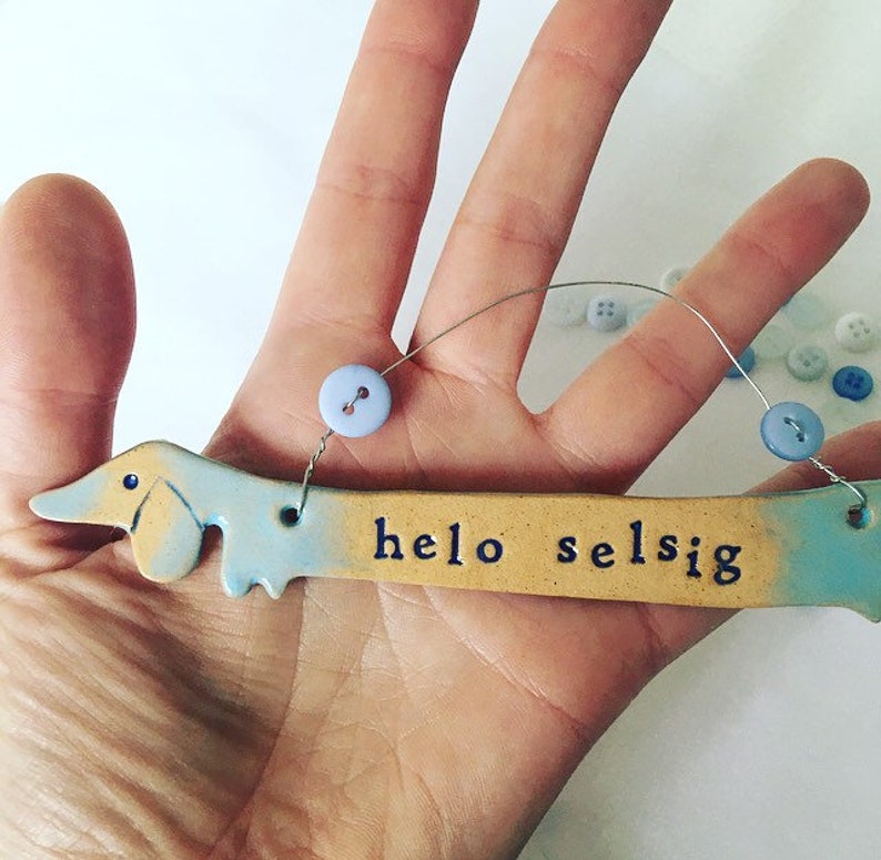 Op de afbeelding: Een keramisch teckelbeeldje met een blauwe en gele glazuur. Het beeldje houdt een bord vast met de tekst "helo selsig".