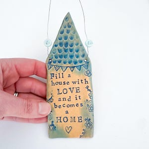 Może przedstawiać: Ceramiczna ozdoba ścienna w kształcie domu z niebieskim dachem w łuski i beżowym korpusem. Tekst głosi: Fill a house with LOVE and it becomes a HOME. Zawiera druciany wieszak z dwoma jasnoniebieskimi koralikami oraz motywami kwiatowymi.