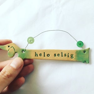 Op de afbeelding: Een keramische dachshund-vormige ornament met een groene en gele glazuur. De ornament heeft een touwtje om op te hangen en staat "hello selsig" op de voorkant.