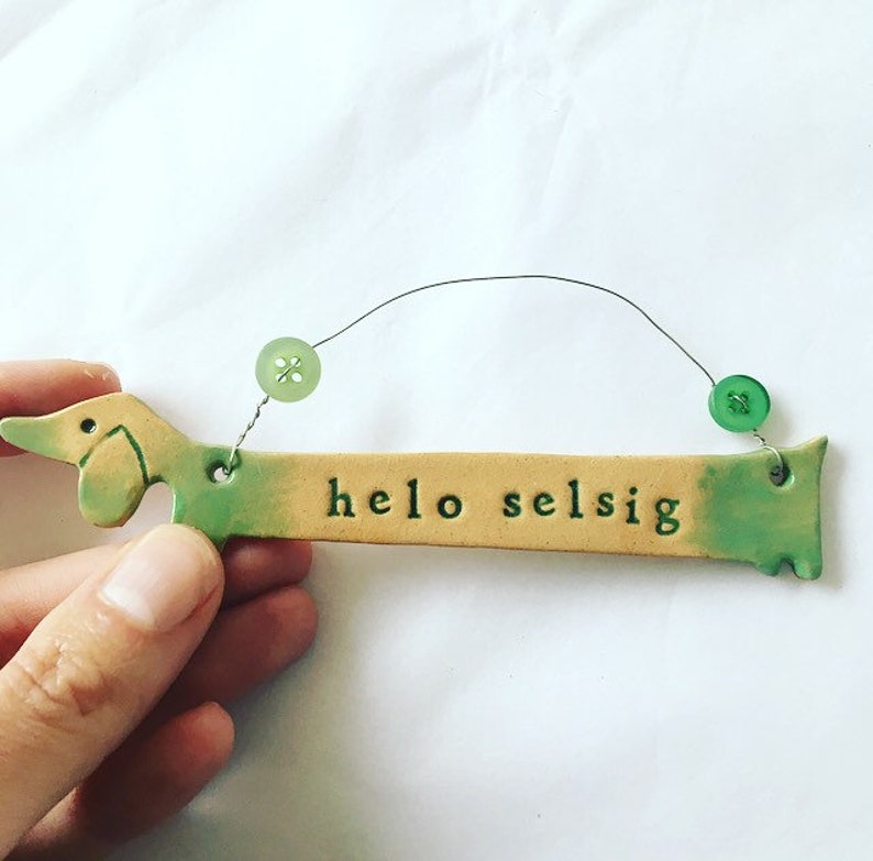 Op de afbeelding: Een keramische dachshund-vormige ornament met een groene en gele glazuur. De ornament heeft een touwtje om op te hangen en staat "hello selsig" op de voorkant.