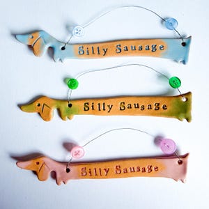 May include: Three ceramic dachshund dog ornaments in shades of blue, green, and pink, each with the words "Silly Sausage" and a button accent. The ornaments are connected by wire.