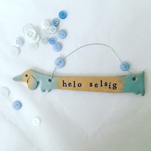 Op de afbeelding: Een keramische dackel-vormige ornament met een blauwe en bruine glazuur. De ornament heeft een draadhanger en de woorden "hello selsig" erop geschreven. De ornament is omringd door blauwe en witte knopen.