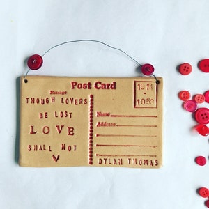 Könnte beinhalten: Eine Keramik-Postkarte mit dem Text "Though lovers be lost love shall not" und einem Herzsymbol. Die Postkarte ist beige mit rotem Text und hat Platz für einen Namen und eine Adresse. Die Postkarte ist datiert 1914-1953 und signiert "Dylan Thomas".