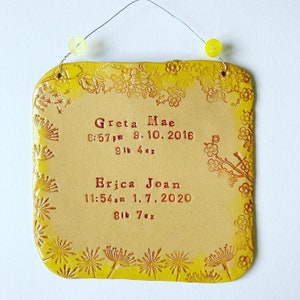 Peut inclure: Une plaque en céramique jaune avec une bordure florale et les noms Greta Mae et Erica Joan, ainsi que leurs dates de naissance et leurs poids. Le texte est en rouge.