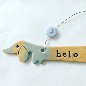 Op de afbeelding: Een keramische dackel-versiering met een blauwe en gele glazuur. De versiering heeft het woord "helo" erop geschreven en hangt aan een draad met een lichtblauwe knoop.