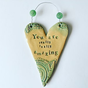Könnte beinhalten: Ein herzförmiger Keramik-Wandbehang mit den Worten "You are really really amazing" in dunkelgrünen Buchstaben. Das Herz ist beige mit grünen dekorativen Mustern und zwei grünen Knöpfen oben.