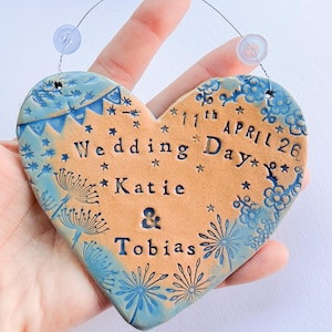 May include: Heart-shaped ceramic wedding plaque with blue floral and banner designs. The plaque has the words "Wedding Day," "Katie & Tobias," and the date "11th April 26" inscribed on it. The plaque is held by a hand.