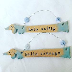 Op de afbeelding: Twee keramische teckel-vormige ornamenten met blauwe en gele glazuur. De bovenste ornament zegt "hello selsig" en de onderste ornament zegt "hello sausage".