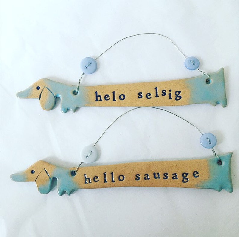 Op de afbeelding: Twee keramische teckel-vormige ornamenten met blauwe en gele glazuur. De bovenste ornament zegt "hello selsig" en de onderste ornament zegt "hello sausage".