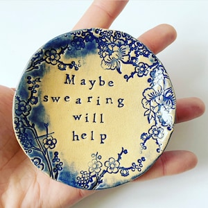 Peut inclure: Petit plat rond en céramique, centre crème et bordure florale bleue. Le texte "Maybe swearing will help" est centré sur le plat en lettres noires. Le plat est tenu dans une main.