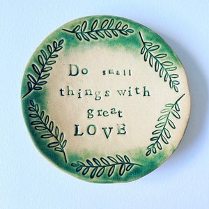 Può includere: Un piatto rotondo in ceramica con un centro color crema e un bordo verde. Il piatto presenta le parole "Do small things with great LOVE" in lettere nere. Un motivo di foglie verdi circonda il bordo del piatto.