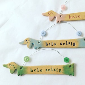 Op de afbeelding: Drie keramische teckel-versieringen met een kleurverloop. Elke versiering heeft een touwtje met een knoop eraan. De versieringen hebben de tekst "hello selsig" erop.