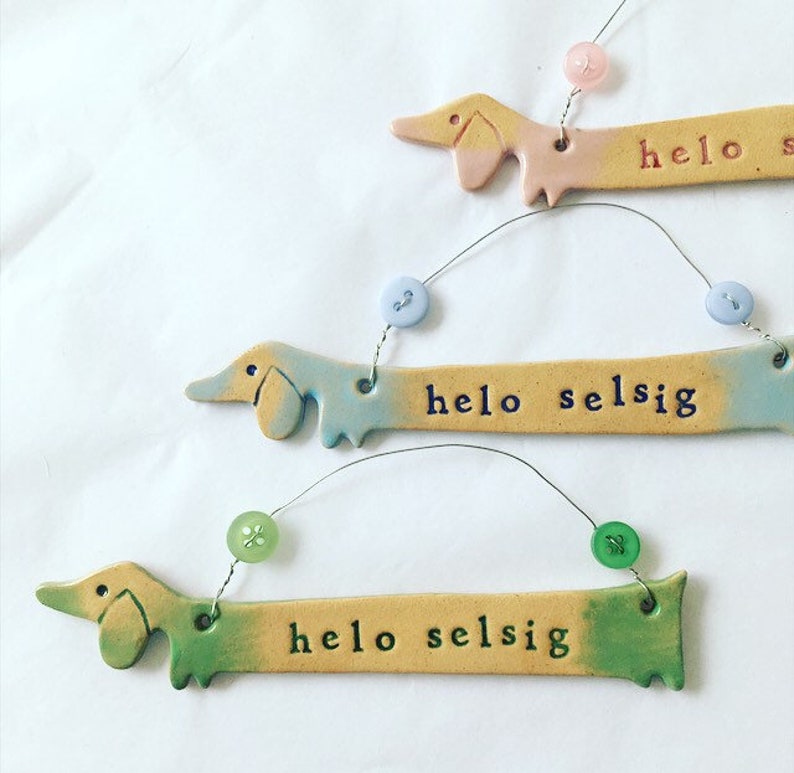 Op de afbeelding: Drie keramische teckel-versieringen met een kleurverloop. Elke versiering heeft een touwtje met een knoop eraan. De versieringen hebben de tekst "hello selsig" erop.